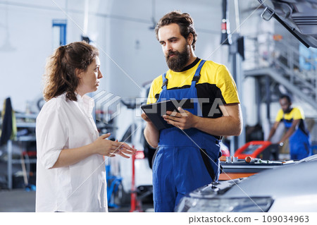 Mechanic in car service ordering new lights system for damaged vehicle using tablet. Worker next to client looking online for components to replace old ones in malfunctioning automobile Mechanic in car service ordering new lights system for damaged vehicle using tablet. Worker next to client looking online for components to replace old ones in malfunctioning automobile 109034963