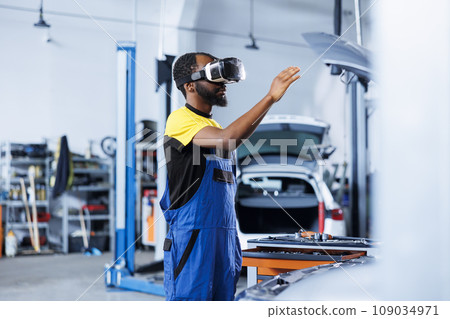 BIPOC repairman in auto repair shop using virtual reality to visualize car battery in order to fix it. Precise garage worker wearing futuristic vr headset while working on busted vehicle BIPOC repairman in auto repair shop using virtual reality to visualize car battery in order to fix it. Precise garage worker wearing futuristic vr headset while working on busted vehicle 109034971