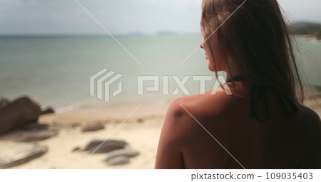 Woman relaxes on balcony on tropical island resort. Luxury spa apartment first line with beautiful seaside view. Back view long hair girl in bikini standing on terrace looking ocean waves. 109035403