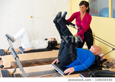 Hispanic woman pilates instructor assisting aged man exercising on reformer Hispanic woman pilates instructor assisting aged man exercising on reformer 109036590