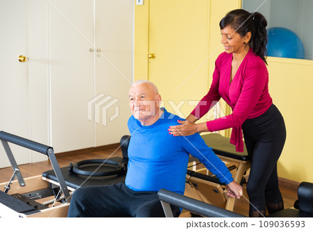 Elderly man practicing pilates on wunda chair under guidance of Hispanic female trainer 109036593