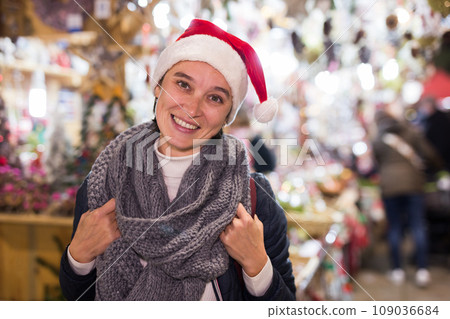 Smiling young woman in Santa hat standing at street Christmas fair Smiling young woman in Santa hat standing at street Christmas fair 109036684