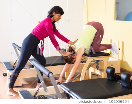 Hispanic woman pilates instructor helping young woman exercising on combo chair 109036994