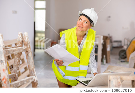 Female foreman in a protective helmet and a yellow vest checks the execution of repair work using laptop 109037010