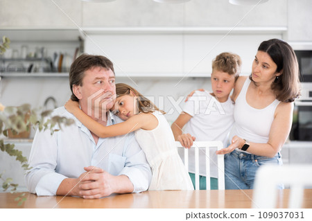 Offended father sitting at the kitchen table being hugged by little daughter Offended father sitting at the kitchen table being hugged by little daughter 109037031