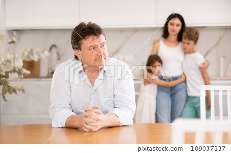 Adult man sitting at table quarreling with family Adult man sitting at table quarreling with family 109037137