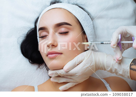 Smile lifting and lip augmentation. Beautician doctor hands doing beauty procedure to female face with syringe. Young woman's mouth contouring with filler injection. Marionette lines treatment Smile lifting and lip augmentation. Beautician doctor hands doing beauty procedure to female face with syringe. Young woman's mouth contouring with filler injection. Marionette lines treatment 109038017