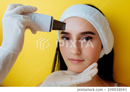 Close up top view of pretty young Caucasian lady during the procedure of cleansing the skin with ultrasound scrubber. Ultrasonic device in the hands of professional female afro beautician. Close up top view of pretty young Caucasian lady during the procedure of cleansing the skin with ultrasound scrubber. Ultrasonic device in the hands of professional female afro beautician. 109038041