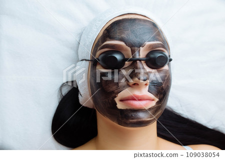 cosmetologist applying black mask on pretty woman face wearing black gloves, gorgeous woman in spa having facial procedures 109038054
