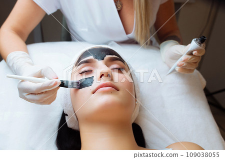 cosmetologist applying black mask on pretty woman face wearing black gloves, gorgeous woman in spa having facial procedures cosmetologist applying black mask on pretty woman face wearing black gloves, gorgeous woman in spa having facial procedures 109038055