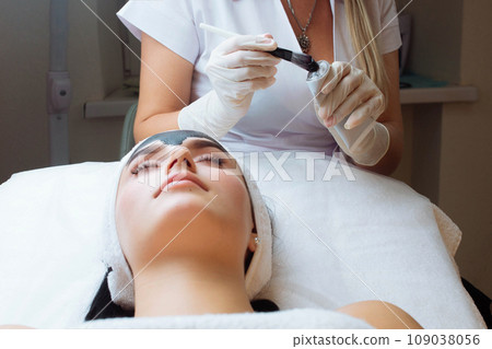 cosmetologist applying black mask on pretty woman face wearing black gloves, gorgeous woman in spa having facial procedures 109038056
