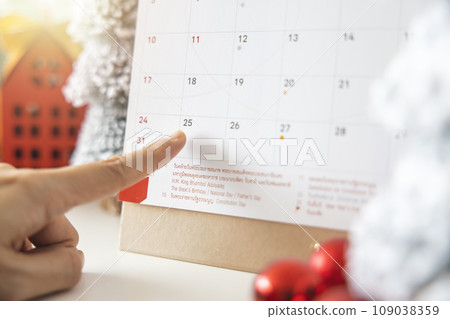 A woman's fingers pointing at the calendar on December 25th, Christmas Day by the festive scene of beautiful Christmas tree adorned, with Christmas balls, pine cones. concept about Important day. 109038359