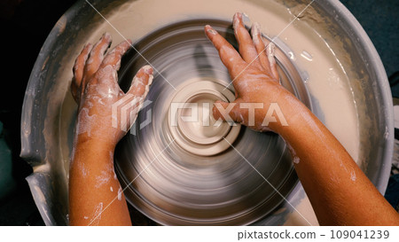 Women's hands stained with clay form a cup blank from clay. View from above. Working on the potter's wheel in the workshop. Close-up. The concept of manual labour, art and craft. 109041239