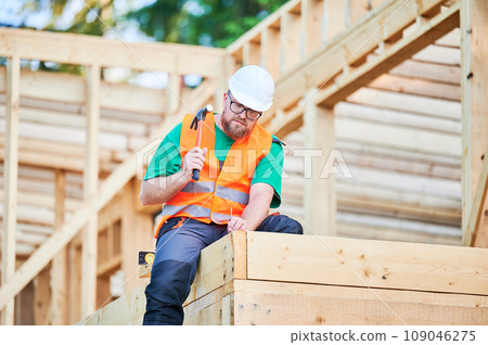 Carpenter constructing wooden frame house. Bearded man wearing glasses hammering nails into structure while wearing helmet and construction vest. Concept of modern ecological construction. 109046275