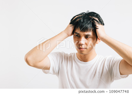 Portrait of a young Asian man with a sad and tired face, holding his head in despair due to stress and a severe headache. Studio shot isolated on white, symbolizing his distress. Portrait of a young Asian man with a sad and tired face, holding his head in despair due to stress and a severe headache. Studio shot isolated on white, symbolizing his distress. 109047748