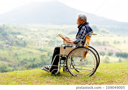 Asian senior man with eyeglasses read the book and sit on wheelchair also stay near the cliff with grassfield and mountain as background. Asian senior man with eyeglasses read the book and sit on wheelchair also stay near the cliff with grassfield and mountain as background. 109049005