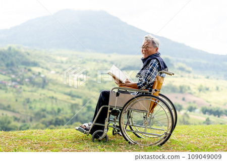 Asian senior man with eyeglasses hold the book and sit on wheelchair also look at camera and stay near the cliff with grassfield and mountain as background. 109049009
