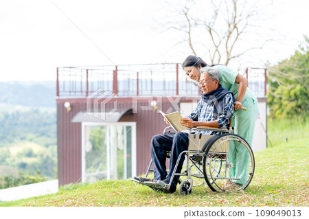 Asian nurse stand behide and look to the book hold by senior man on wheelchair and both look happy and stay near cliff with village and mountain in the background. 109049013