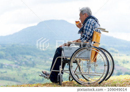 Asian senior man drink tea from a cup and sit on wheelchair with near the cliff and mountain and grassfield as background. 109049014
