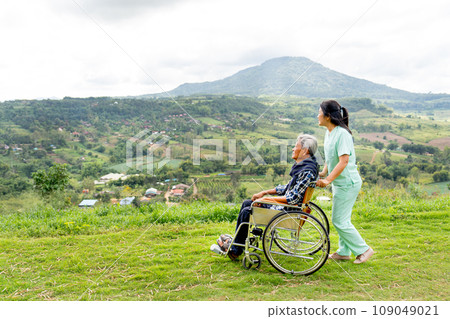 Asian nurse stand and hold wheelchair hand that sit by senior man and both stay near cliff with village and mountain in the background. Asian nurse stand and hold wheelchair hand that sit by senior man and both stay near cliff with village and mountain in the background. 109049021