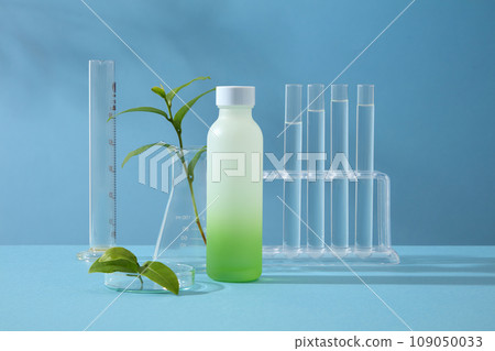Mockup scene for advertising cosmetic of green tea extract. An empty bottle displayed with lab glassware and tea bud on blue background. Mockup for design, organic cosmetics, beauty products concept 109050033
