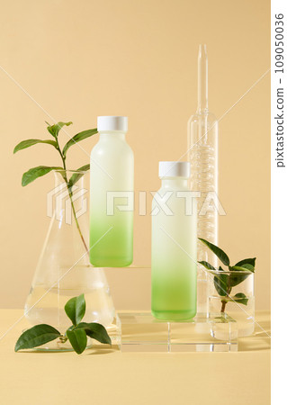 Set of cosmetic bottles on glass podiums with green tea leaves and lab glassware on beige background. Product and promotion concept for advertising cosmetic of green tea extract. Front view  109050036
