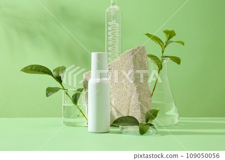 Mockup scene for advertising cosmetic of natural extract - green tea. On green background, cosmetic bottle mockup displayed with fresh leaves and bud of green tea, stone and lab glassware. Front view 109050056