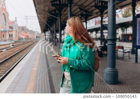 Young redhead woman waiting train with backpack and using smart phone. Railroad transport concept, Traveler. 30s Woman walking at railroad station platform. Travel to vacation by train 109050143