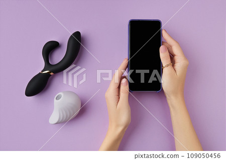 Top view of female hands holding a cellphone with adult sex toys on the purple background Top view of female hands holding a cellphone with adult sex toys on the purple background 109050456