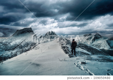 Mountaineer standing on top of snowy mountain with dark stormy sky in winter on Segla mount at Senja Island Mountaineer standing on top of snowy mountain with dark stormy sky in winter on Segla mount at Senja Island 109050480