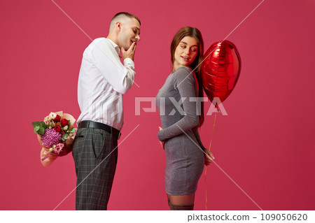 Holiday. Man hiding flowers behind his back for girlfriend, girl holding balloon against pink studio background. Valentine's day, relationship 109050620