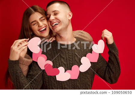 Happy, caring, loving young couple, boy and girl holding paper heart garland, celebrating holiday against red studio background. Valentine's day, relationship Happy, caring, loving young couple, boy and girl holding paper heart garland, celebrating holiday against red studio background. Valentine's day, relationship 109050650