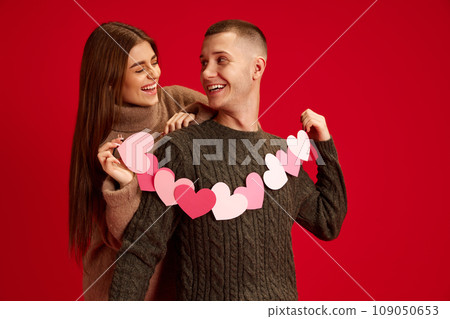 Celebrating holidays. Young happy loving couple, man and woman holding hearts, posing against red studio background. Valentine's day, relationship Celebrating holidays. Young happy loving couple, man and woman holding hearts, posing against red studio background. Valentine's day, relationship 109050653