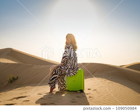 Arabian woman in stylish zebra suit with bright light green or yellow suitcase in sands. Concept and idea of travel to United Arab Emirates, sand dunes in desert of Dubai at sunset. 109050984
