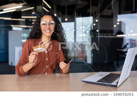 Portrait of business woman inside office at workplace, Hispanic woman smiling looking at camera, working with laptop, female worker holding phone and bank debit credit card, shopping online. 109059201