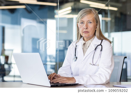 Portrait of a senior gray-haired female doctor sitting in the office, working online on a laptop and looking seriously into the camera. 109059202