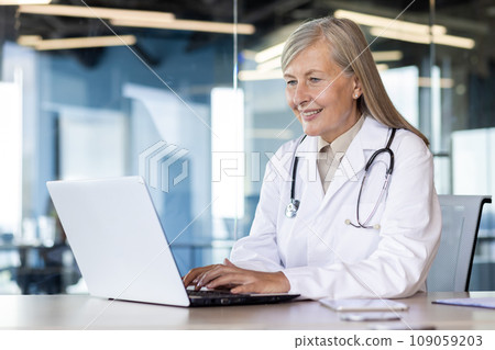 Senior gray-haired female doctor consults and talks online with patients via video call from laptop. Sits at the table and works in the office of the clinic. 109059203