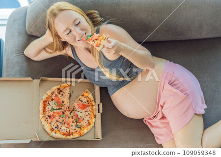 A pregnant woman enjoys a slice of pizza, savoring a moment of indulgence while satisfying her craving for a delightful, comforting treat. Excited Pregnant Young Lady Enjoying Pizza Holding Biting 109059348