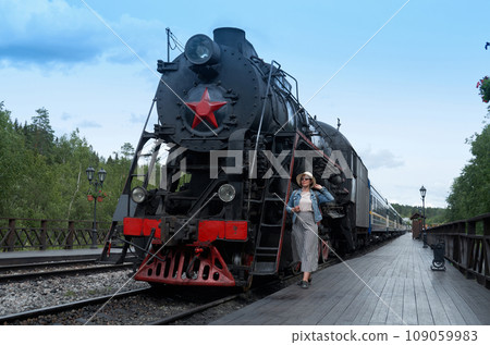A female tourist in a straw hat and denim jacket stands on the platforms next to a steam train 109059983
