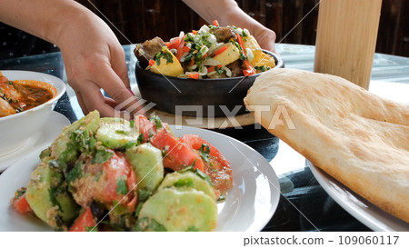 Close-up of waitress putting Ojahuri (dish made of stewed meat (pork) with potatoes, onions and bell peppers) and grape lemonade on a table on table. Georgian traditional food concept Close-up of waitress putting Ojahuri (dish made of stewed meat (pork) with potatoes, onions and bell peppers) and grape lemonade on a table on table. Georgian traditional food concept 109060117