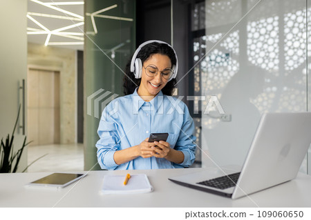 Young beautiful woman at workplace joyful listening to music and online radio, using app on phone, businesswoman resting on break, singing along well done job well done. Young beautiful woman at workplace joyful listening to music and online radio, using app on phone, businesswoman resting on break, singing along well done job well done. 109060650