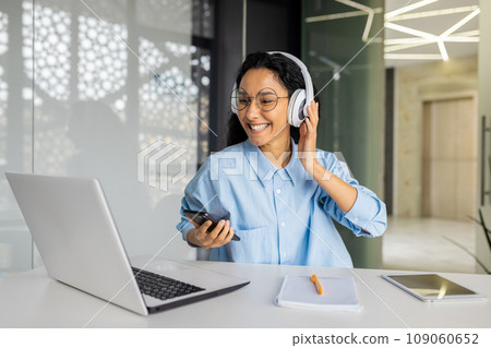 Young beautiful woman at workplace joyful listening to music and online radio, using app on phone, businesswoman resting on break, singing along well done job well done. Young beautiful woman at workplace joyful listening to music and online radio, using app on phone, businesswoman resting on break, singing along well done job well done. 109060652