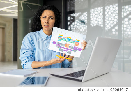 Portrait of dissatisfied excited businesswoman at workplace inside office, female worker with laptop looking disappointed, upset at camera, showing table report with negative results of achievements. Portrait of dissatisfied excited businesswoman at workplace inside office, female worker with laptop looking disappointed, upset at camera, showing table report with negative results of achievements. 109061647