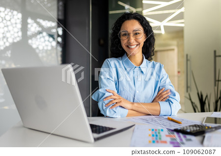 Portrait of young successful female accountant accountant at workplace, satisfied hispanic woman with crossed arms looking at camera while sitting inside office at workplace with laptop. Portrait of young successful female accountant accountant at workplace, satisfied hispanic woman with crossed arms looking at camera while sitting inside office at workplace with laptop. 109062087