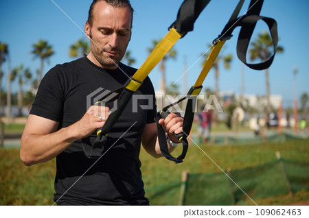 Determined muscular build young adult man, sportsman, athlete exercising with suspension straps outdoors 109062463