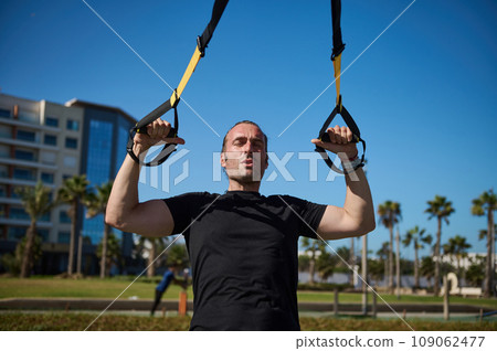 Caucasian middle aged athletic man in black t-shirt exercising with suspension fitness straps in urban background Caucasian middle aged athletic man in black t-shirt exercising with suspension fitness straps in urban background 109062477