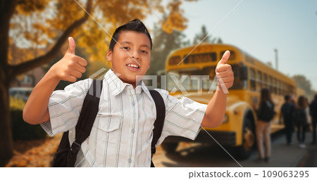 Excited Young Hispanic Boy Wearing a Backpack Giving Two Thumbs Up on Campus Near a School Bus 109063295