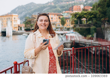 Hipster girl with two tickets browsing Internet. Cruise ship vacation concept. Varenna, Italy. Lake Como. Phone Communication. Female holding phone and booking hotel Hipster girl with two tickets browsing Internet. Cruise ship vacation concept. Varenna, Italy. Lake Como. Phone Communication. Female holding phone and booking hotel 109063369