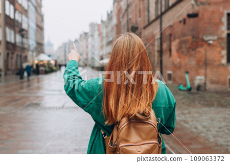 Casual dressed woman 30 years old exploring city streets during international excursion in town. Redhead girl with backpack pointing finger on city street in Gdansk. Traveling Europe in autumn. Casual dressed woman 30 years old exploring city streets during international excursion in town. Redhead girl with backpack pointing finger on city street in Gdansk. Traveling Europe in autumn. 109063372