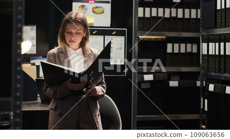 Female private detective conducting investigation in office, focused on reviewing files and evidence. Portrait shot of policewoman analyzing statements and records. Looking at camera, tripod shot. 109063656